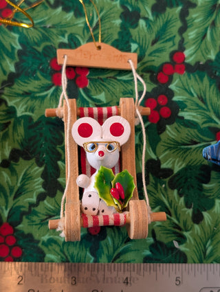 Decorative Christmas ornament of a mouse on a swing with a green leaf, against a festive background.